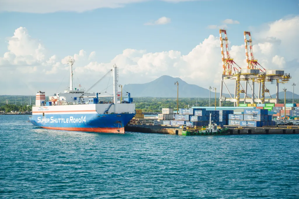 A cargo ship docked at an industrial port with shipping containers and cranes under a clear sky.