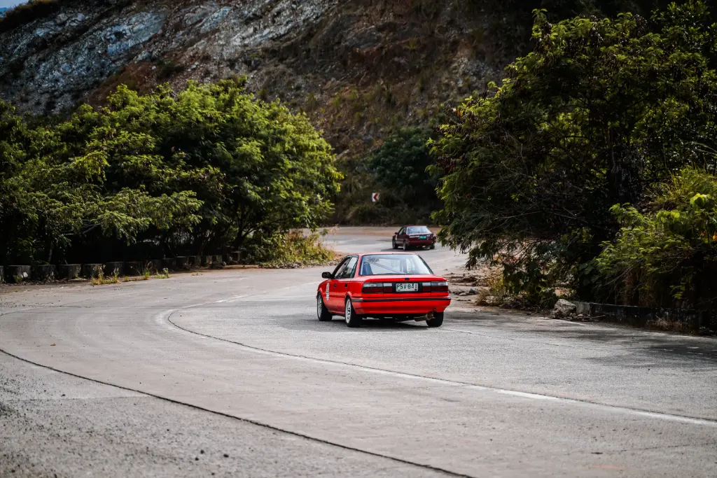 A red car driving on a scenic mountain road in Mangatarem, Philippines, surrounded by greenery and hills.