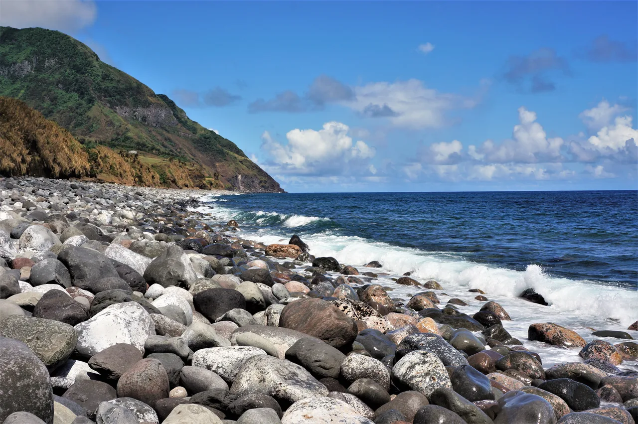 Große Steine und Brandung am Valugan Boulder Beach vor einer grünen Küste