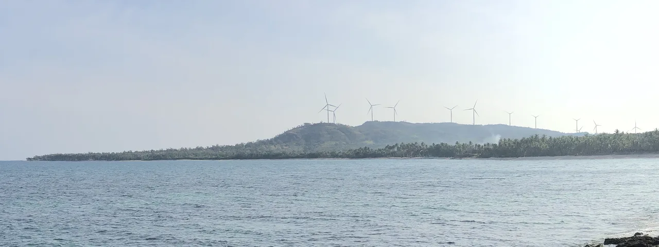 Blick von Saud Beach über das Meer auf eine palmenreiche Küste und die Windturbinen von Caparispisan