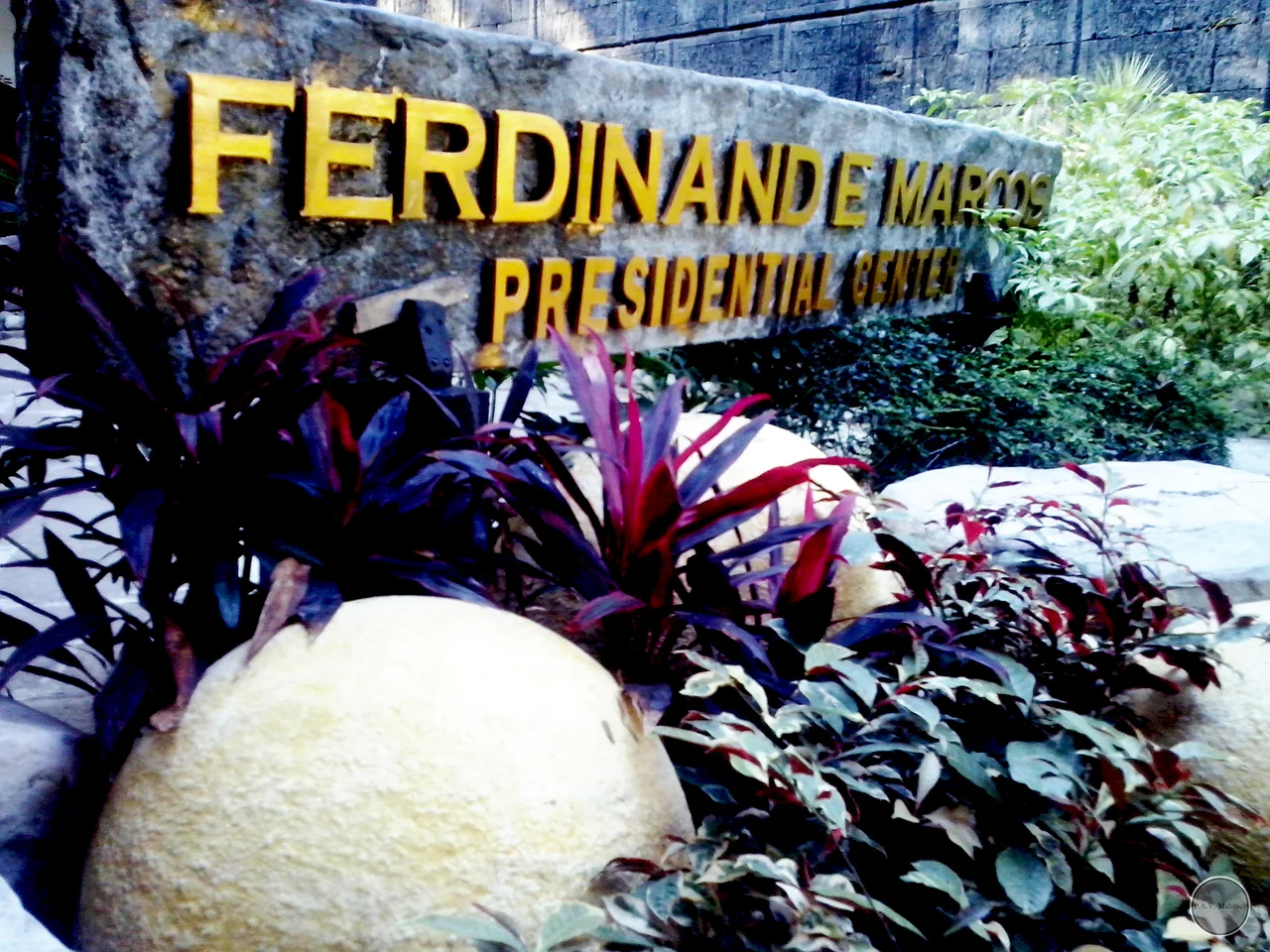 Steinernes Namensschild des Ferdinand E. Marcos Presidential Center mit gelben Buchstaben zwischen Pflanzen
