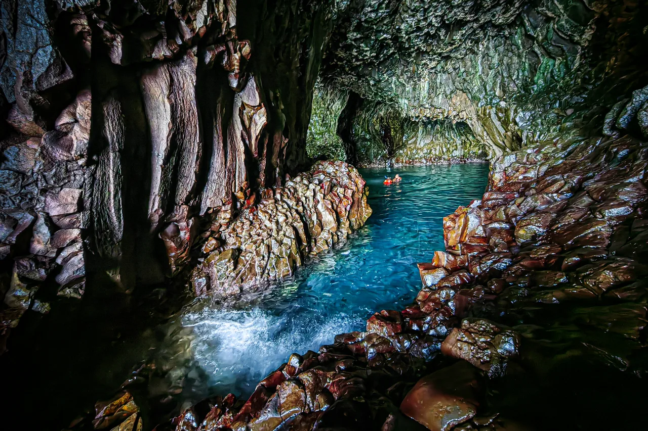 Felsige Küstenhöhle mit blaugrünem Wasser und kleinen Schwimmern im Hintergrund